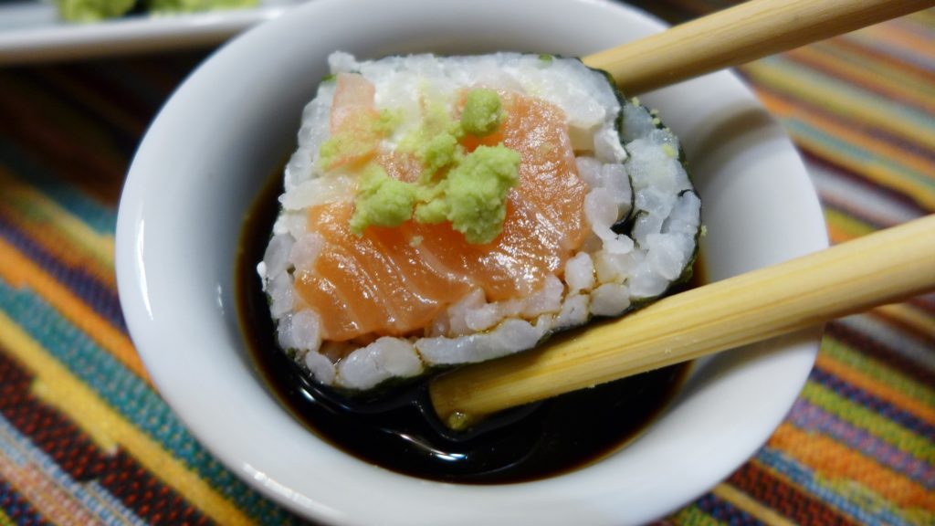 Ein Maki-Sushi mit Wasabi wird in ein Schälchen mit Sojasoße getunkt.