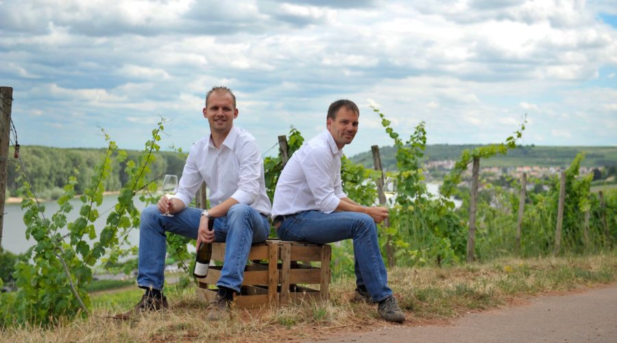 Winzer Stefan und Daniel Huff vom Weingut Georg Gustav Huff sitzen auf Weinkisten vor ihrem Weinberg und trinken Wein