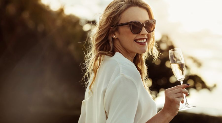 Elegante Frau mit blonden Haaren und Sonnenbrille im Sonnenuntergang mit einem Glas Crémant in der Hand