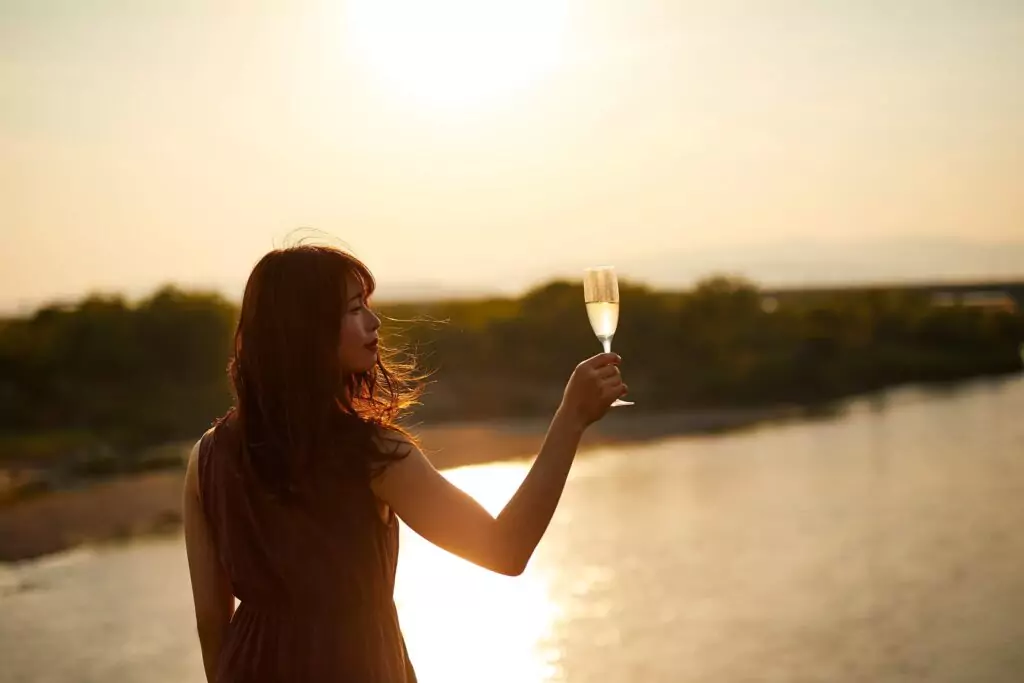 Frau an einem Fluss bei Sonnenuntergang mit einem Glas Crémant de Bordeaux in der Hand