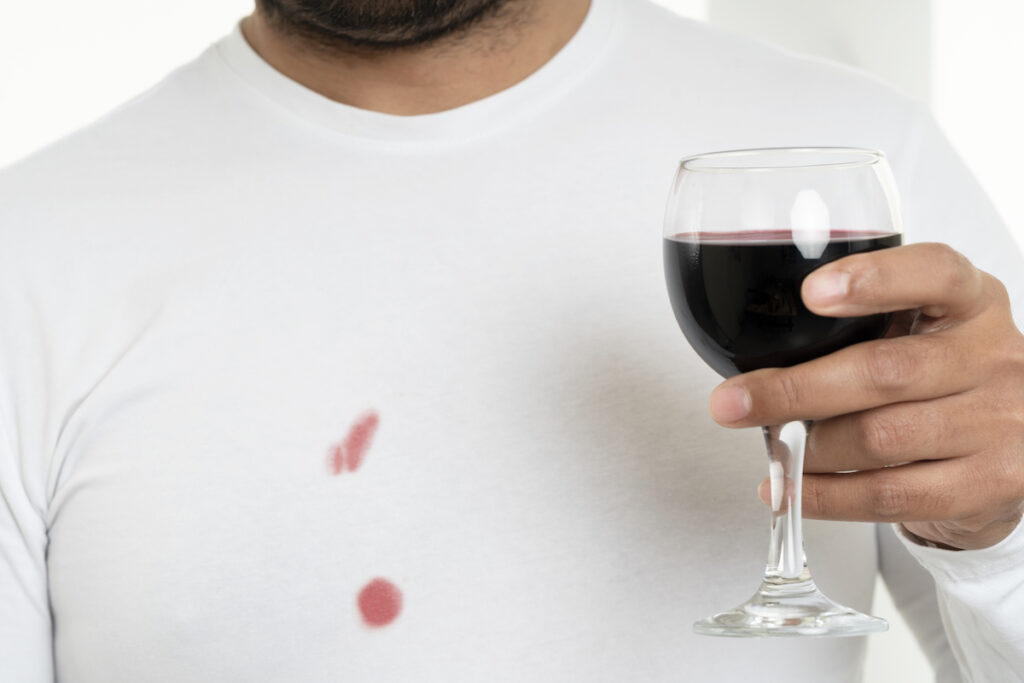 Mann hat Rotweinflecken auf seinem weißen T-Shirt und hält ein Rotweinglas in der Hand