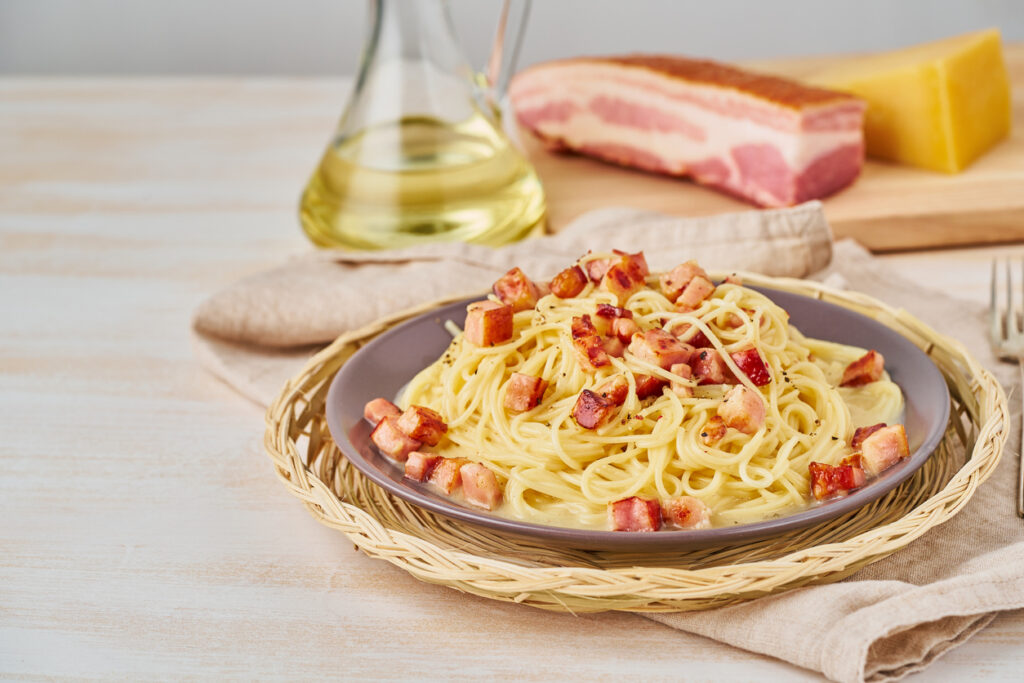 Grauer Teller mit Spaghetti Carbonara - im Hintergrund sind die Zutaten zu sehen